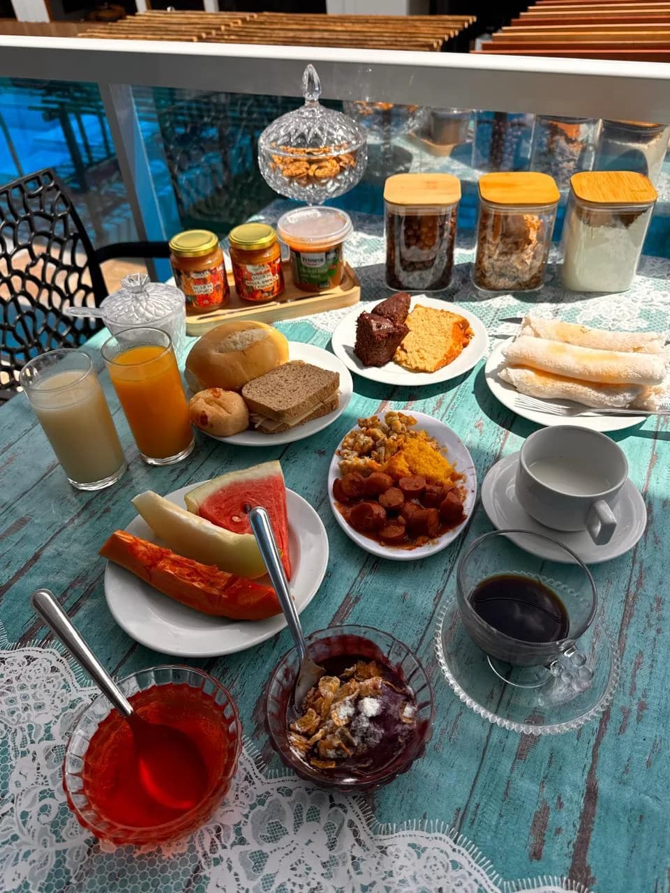 Mesa do café da manhã com tapiocas, cuscuz com calabresa, frutas, bolo, pães, sucos e café, ao lado da piscina