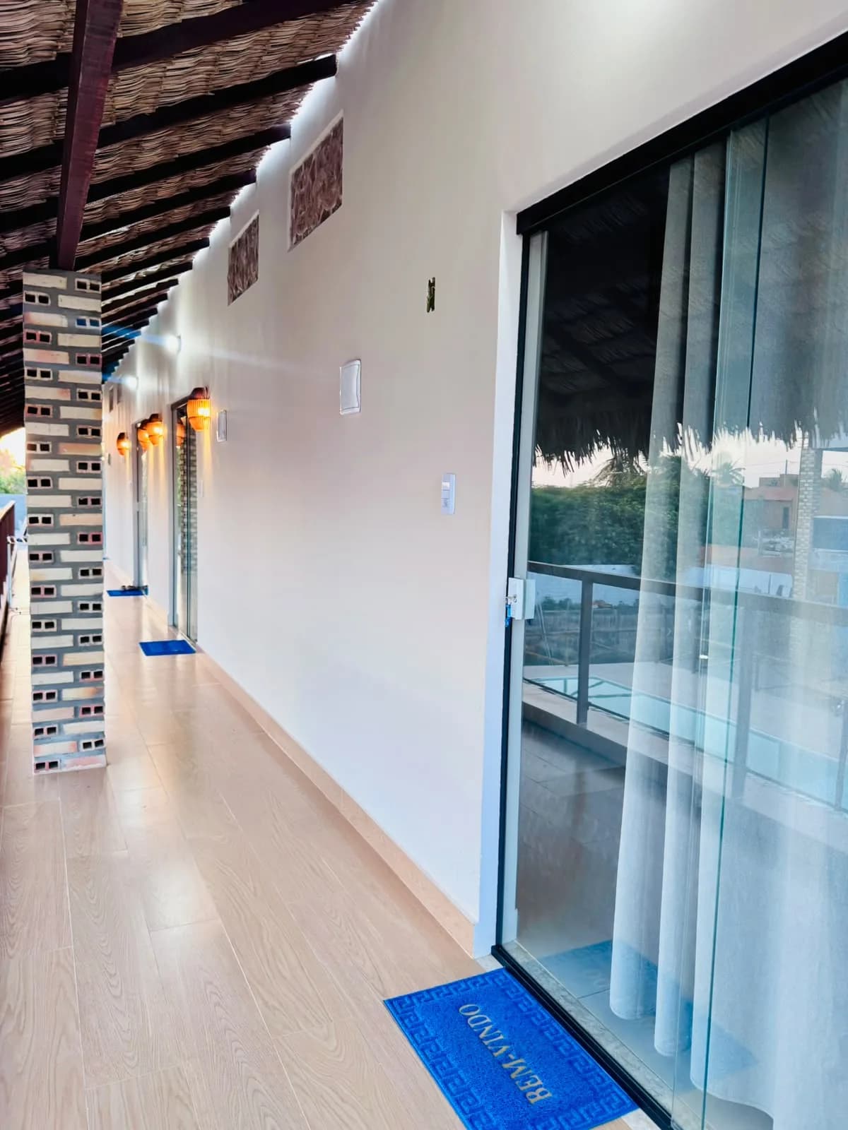 Corredor dos quartos com cobertura de palha, piso amadeirado, portas de vidro e tapete de boas-vindas