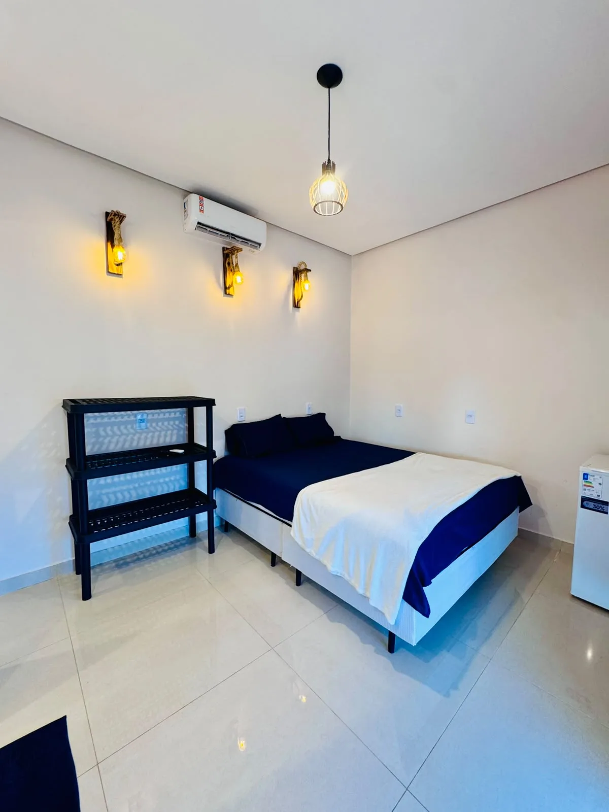 Quarto com cama de casal de lençóis azuis, ar-condicionado, frigobar e arandelas de luz amarela