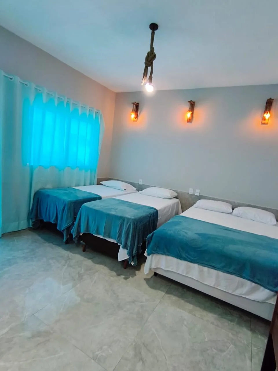 Quarto amplo com três camas, roupa de cama branca com mantas azuis, cortina turquesa e arandelas de luz amarela